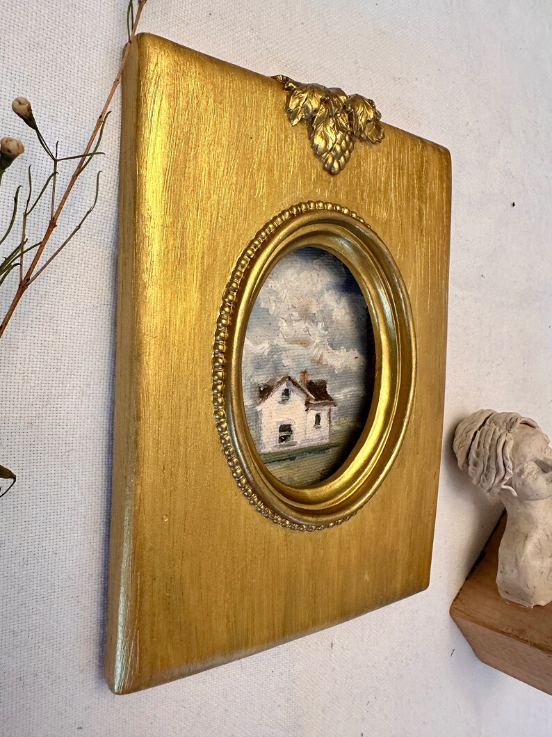 May include: A rectangular gold-painted frame with a central oval opening featuring a beaded inner edge and a decorative leaf motif at the top. The frame contains a small oil painting of a white house under a cloudy sky with visible brushstrokes.
