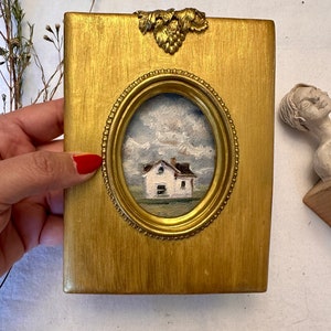 May include: A rectangular gold-painted wooden frame featuring an oval center cutout that displays a small oil painting of a white house under a cloudy blue sky. The frame is adorned with a decorative metallic grape leaf accent at the top center.