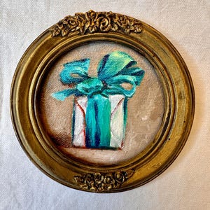 May include: A small circular painting of a white gift box wrapped with a vibrant teal ribbon and bow. The artwork is encased in an ornate, antique gold-toned frame featuring detailed floral carvings at the top and bottom edges of the circular border.