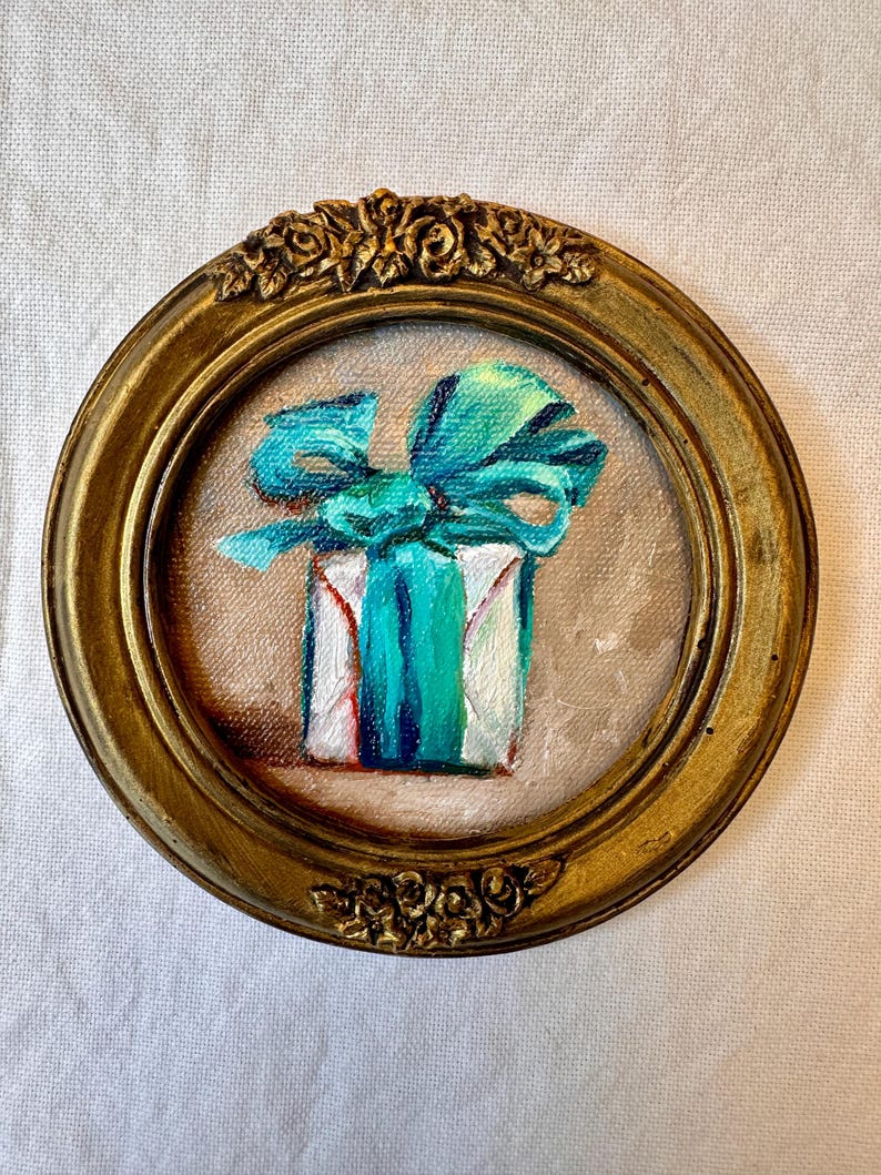 May include: A small circular painting of a white gift box wrapped with a vibrant teal ribbon and bow. The artwork is encased in an ornate, antique gold-toned frame featuring detailed floral carvings at the top and bottom edges of the circular border.