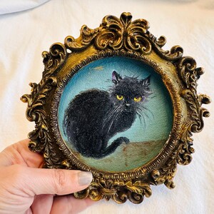 May include: A small, round painting of a black cat with yellow eyes, set in an ornate, antique gold-colored frame. The cat is depicted against a light blue background. The frame has intricate scrollwork.