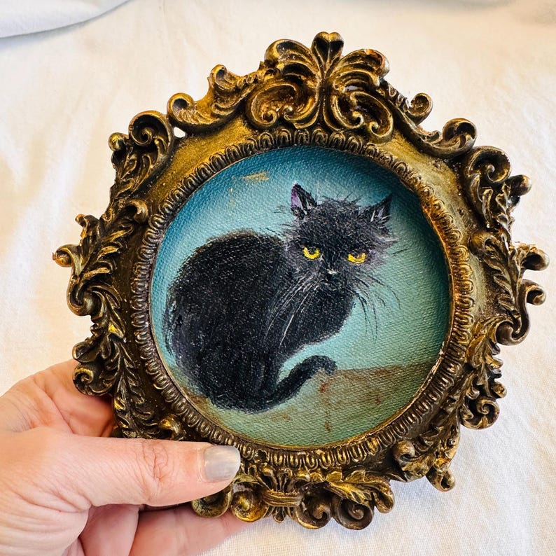 May include: A small, round painting of a black cat with yellow eyes, set in an ornate, antique gold-colored frame. The cat is depicted against a light blue background. The frame has intricate scrollwork.