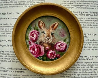 May include: A small circular painting of a brown rabbit surrounded by pink roses, encased in a textured gold-colored frame. The artwork features detailed brushstrokes depicting the rabbit's fur and the layered petals of the vibrant pink flowers.