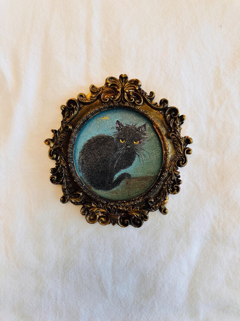 May include: A vintage-style framed artwork featuring a black cat with yellow eyes against a blue background. The cat is depicted in a seated position. The ornate, oval frame has a gold finish, adding a touch of antique charm to the piece.