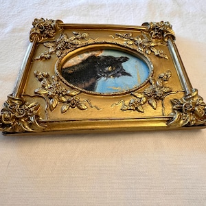 K&ouml;nnte beinhalten: Ein Portr&auml;tgem&auml;lde einer schwarzen Katze in einem goldenen, verzierten Rahmen mit floralen Details. Das Gem&auml;lde ist oval geformt und die Katze schaut nach rechts.
