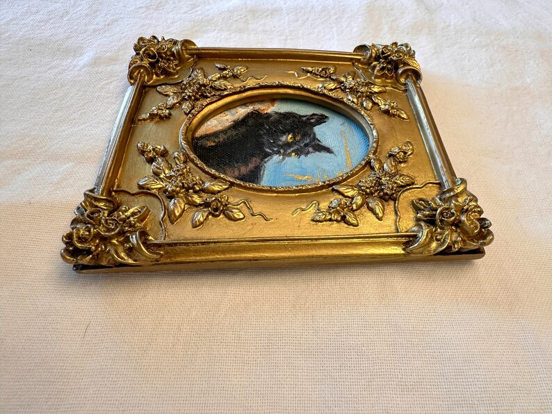 K&ouml;nnte beinhalten: Ein Portr&auml;tgem&auml;lde einer schwarzen Katze in einem goldenen, verzierten Rahmen mit floralen Details. Das Gem&auml;lde ist oval geformt und die Katze schaut nach rechts.