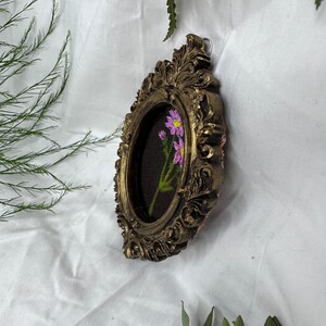 May include: A small oval-shaped picture frame with ornate gold detailing. The frame contains a hand-painted floral design with pink flowers on a dark green background.