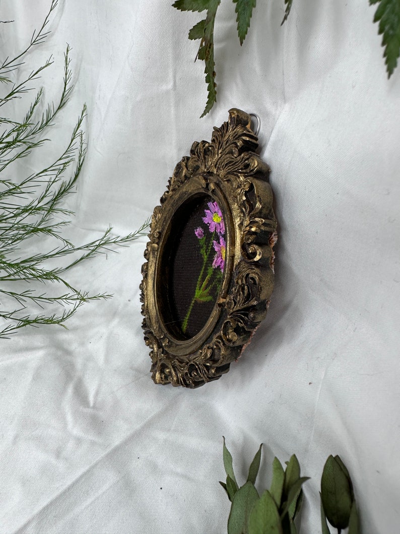 May include: A small oval-shaped picture frame with ornate gold detailing. The frame contains a hand-painted floral design with pink flowers on a dark green background.