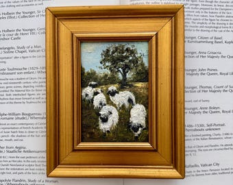 May include: A small oil painting in a gold-toned frame depicting a group of five white sheep with black faces and legs standing in a grassy field under a single tree. The painting features visible brushstrokes and a textured application of paint.