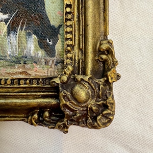 May include: A framed painting featuring a black and white cow grazing in a field. The frame is ornate, finished in a metallic gold color with raised, decorative scrollwork details at the corners and along the edges of the rectangular border.