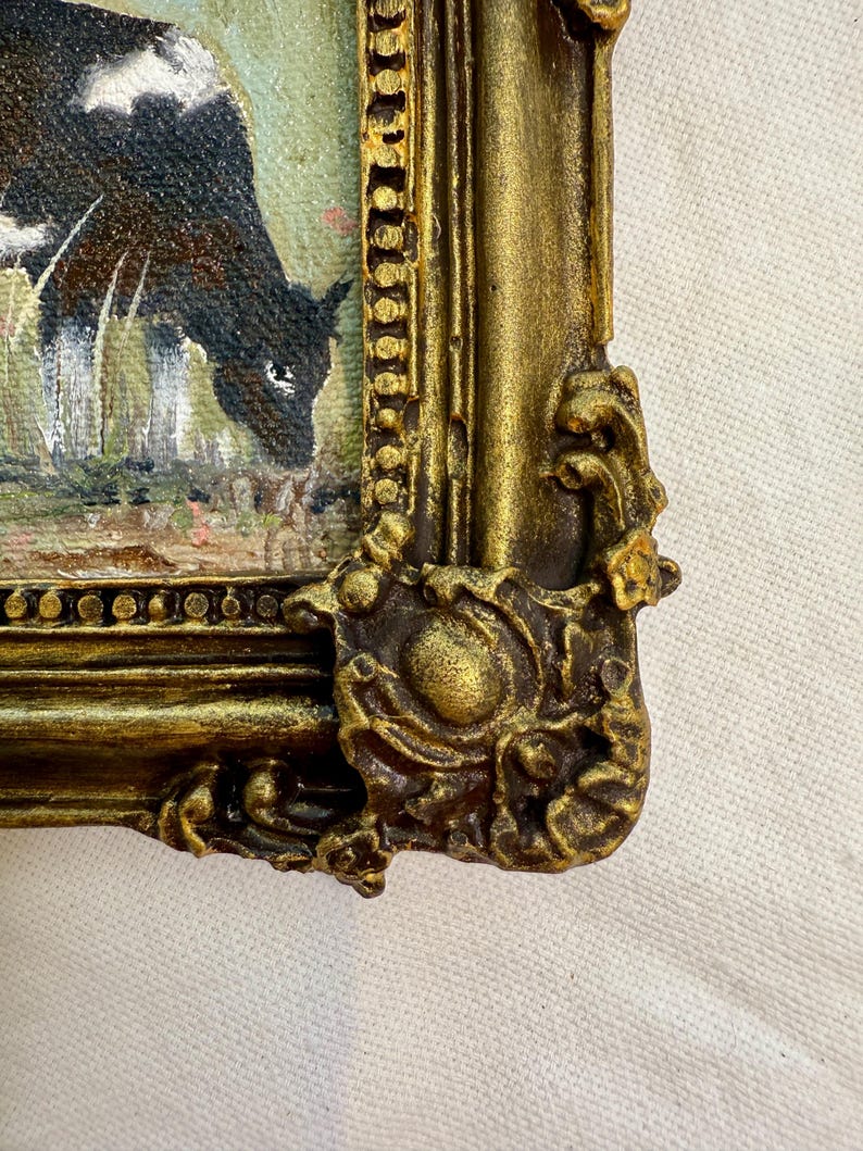 May include: A framed painting featuring a black and white cow grazing in a field. The frame is ornate, finished in a metallic gold color with raised, decorative scrollwork details at the corners and along the edges of the rectangular border.