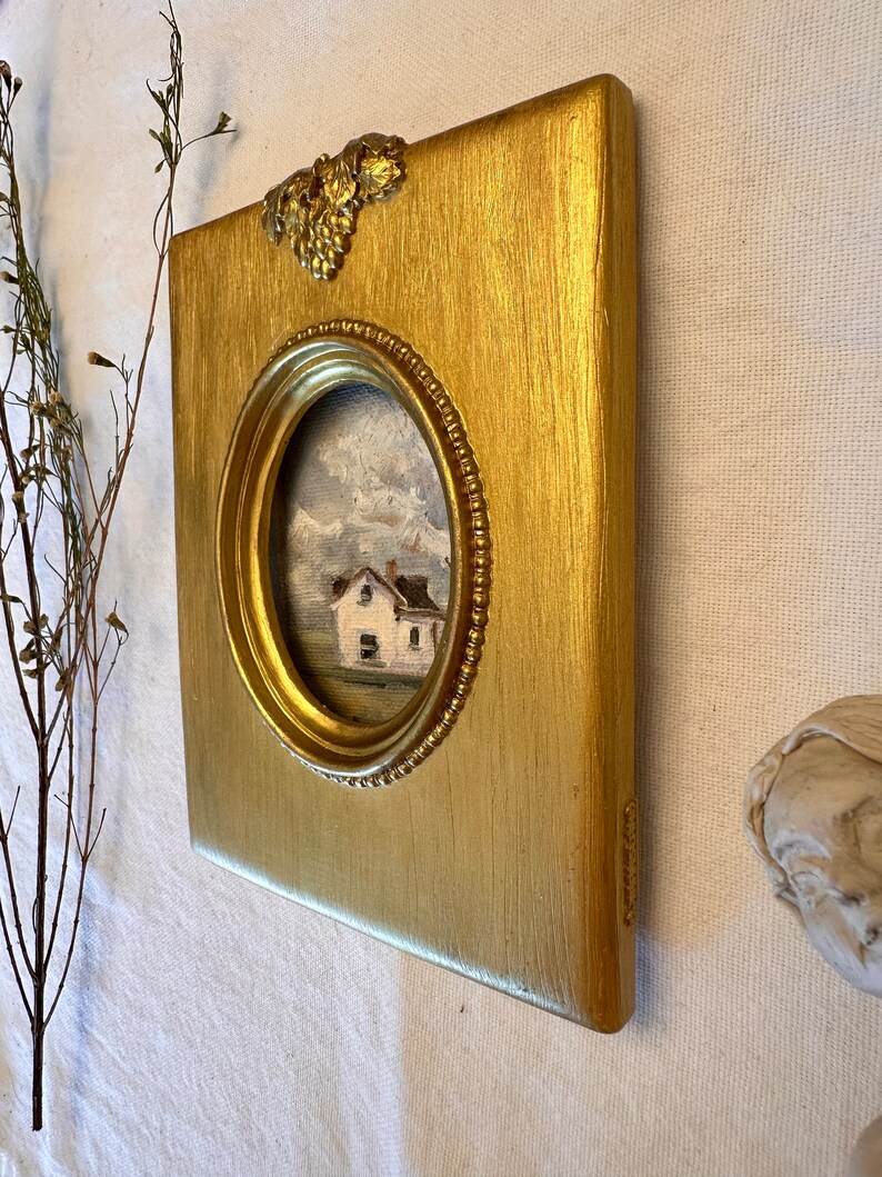 May include: A rectangular gold-painted frame with a textured finish and a decorative grape cluster accent at the top. The frame features a central oval opening containing a small oil painting of a white house set against a cloudy, mountainous landscape.