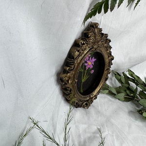 May include: A small, oval-shaped, gold-colored picture frame with ornate detailing. The frame contains an embroidered image of a single pink flower with green leaves and stems on a black background.