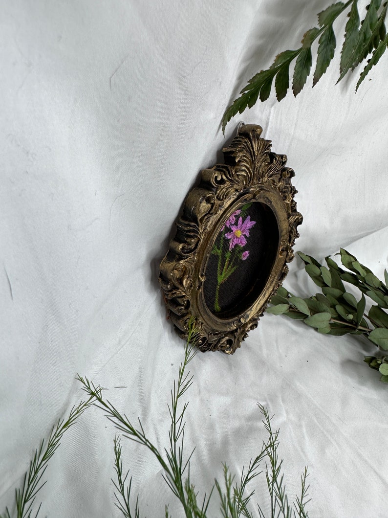 May include: A small, oval-shaped, gold-colored picture frame with ornate detailing. The frame contains an embroidered image of a single pink flower with green leaves and stems on a black background.
