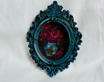 May include: An oval wall frame with a dark teal, distressed finish and ornate, scrolling relief details. The center contains a textured, dark-toned needlepoint or cross-stitch artwork depicting a small blue vase filled with vibrant red flowers.
