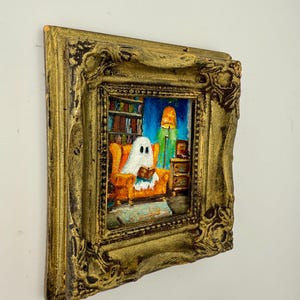 May include: A small, framed painting depicts a ghost reading in an orange armchair. The artwork is set in a room with a bookshelf, a lamp, and a side table. The frame is ornate and gold-colored, adding a vintage touch to the artwork.