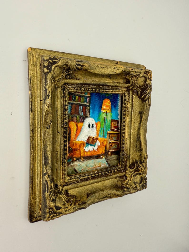 May include: A small, framed painting depicts a ghost reading in an orange armchair. The artwork is set in a room with a bookshelf, a lamp, and a side table. The frame is ornate and gold-colored, adding a vintage touch to the artwork.
