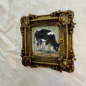 May include: A small square painting of a black and white cow grazing in a field, enclosed in an ornate, heavy gold-colored frame with intricate relief detailing along the edges and corners, featuring a textured finish and a deep, metallic luster.