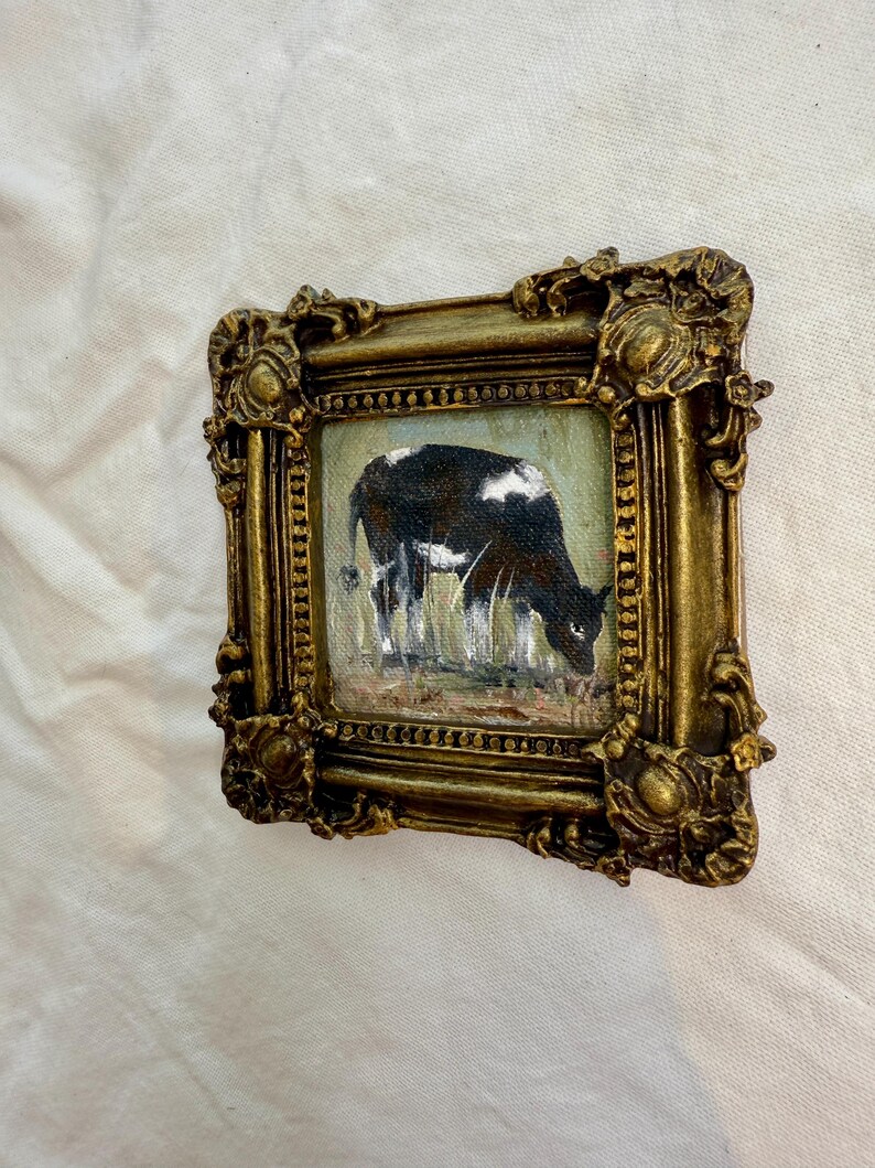 May include: A small square painting of a black and white cow grazing in a field, enclosed in an ornate, heavy gold-colored frame with intricate relief detailing along the edges and corners, featuring a textured finish and a deep, metallic luster.