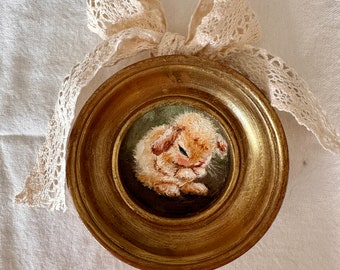 May include: A small, round, gold-framed picture of a white bunny rabbit with brown eyes. The bunny is sitting on a green background and is looking down. The frame is decorated with a white lace bow.