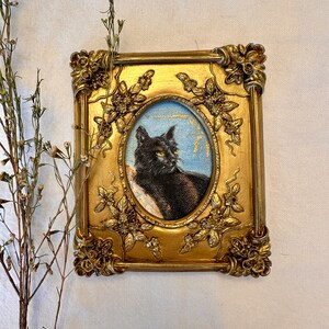 K&ouml;nnte beinhalten: Ein gerahmtes Gem&auml;lde einer schwarzen Katze mit gelben Augen. Die Katze sitzt auf einem blauen Hintergrund mit goldenen Akzenten. Der Rahmen ist golden mit verzierten Details.