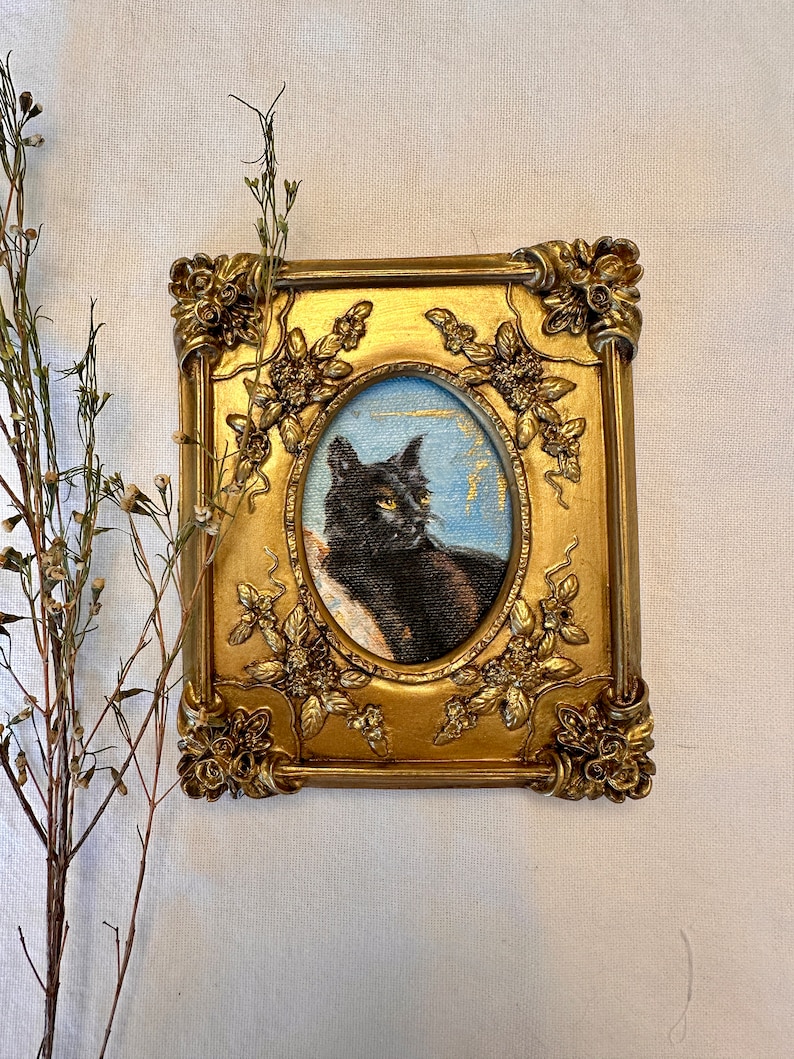 K&ouml;nnte beinhalten: Ein gerahmtes Gem&auml;lde einer schwarzen Katze mit gelben Augen. Die Katze sitzt auf einem blauen Hintergrund mit goldenen Akzenten. Der Rahmen ist golden mit verzierten Details.