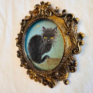 May include: A framed painting of a black cat with yellow eyes. The cat is depicted against a light blue background, within an ornate, gold-colored oval frame. The frame has detailed scrollwork and a vintage aesthetic.