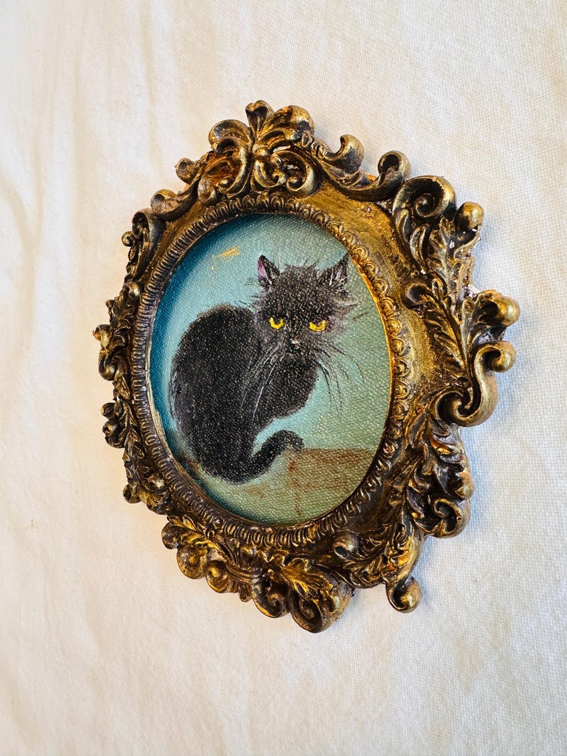 May include: A framed painting of a black cat with yellow eyes. The cat is depicted against a light blue background, within an ornate, gold-colored oval frame. The frame has detailed scrollwork and a vintage aesthetic.