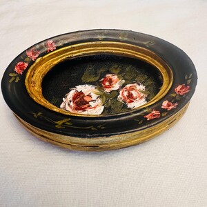 May include: An oval-shaped trinket dish with a black base and gold rim. The dish is decorated with hand-painted pink roses and green leaves.