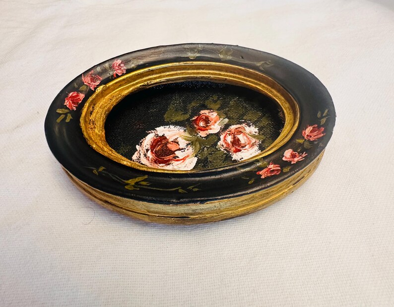May include: An oval-shaped trinket dish with a black base and gold rim. The dish is decorated with hand-painted pink roses and green leaves.