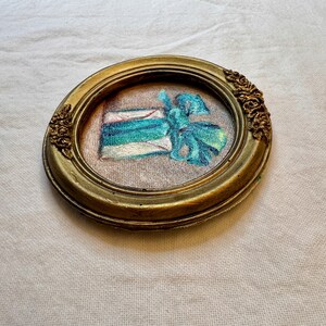 May include: An oval-shaped gold-toned frame with ornate floral detailing on the sides. Inside the frame is a piece of fabric featuring an embroidered design of a gift box wrapped in teal and blue ribbon with a prominent bow on the right side.