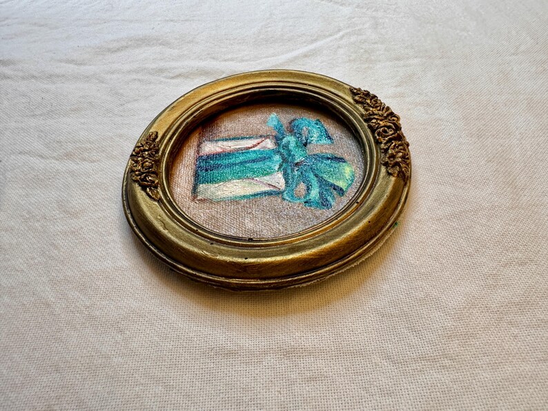 May include: An oval-shaped gold-toned frame with ornate floral detailing on the sides. Inside the frame is a piece of fabric featuring an embroidered design of a gift box wrapped in teal and blue ribbon with a prominent bow on the right side.