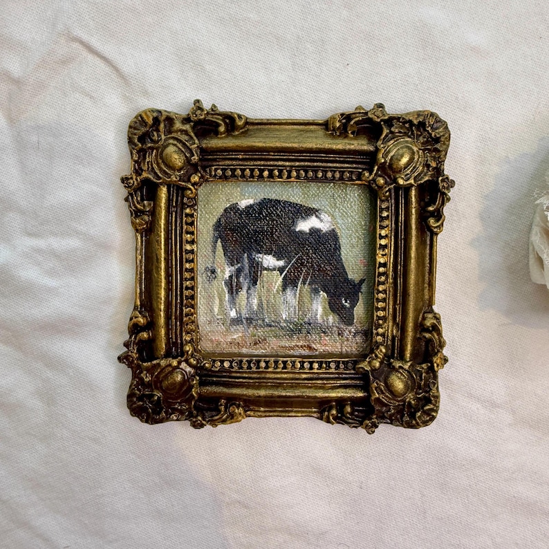 May include: A small square painting of a black and white cow grazing in a field, enclosed within an ornate, antique-style gold frame featuring intricate scrollwork and raised decorative details along the edges of the heavy metallic border.