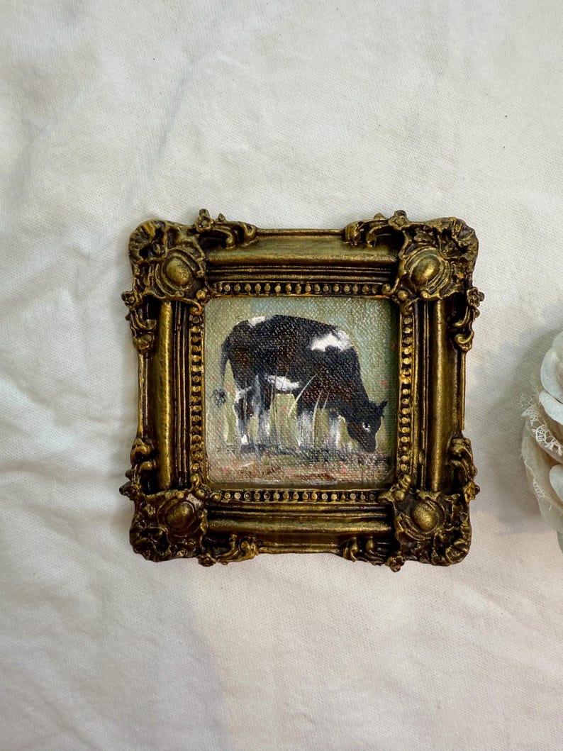 May include: A small square painting of a black and white cow grazing in a field, enclosed in an ornate, antique-style gold frame with intricate scrollwork and bead detailing along the inner border, showcasing a textured, painterly finish.
