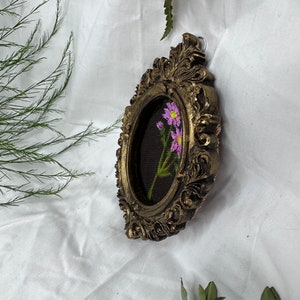 May include: A small oval-shaped picture frame with ornate gold detailing. The frame contains a black background with a single stem of pink flowers embroidered in the center.