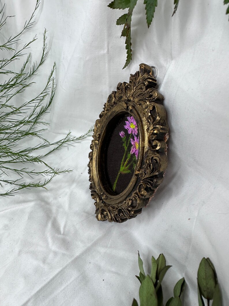 May include: A small oval-shaped picture frame with ornate gold detailing. The frame contains a black background with a single stem of pink flowers embroidered in the center.