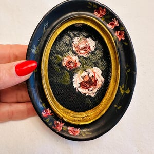 May include: A small oval-shaped framed painting of pink roses on a black background. The frame is black with a gold inner border and has pink roses painted around the edge.