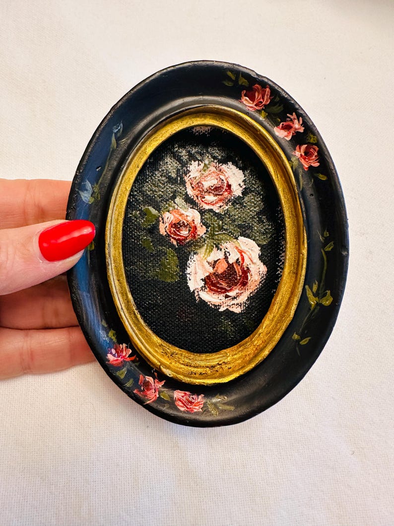 May include: A small oval-shaped framed painting of pink roses on a black background. The frame is black with a gold inner border and has pink roses painted around the edge.