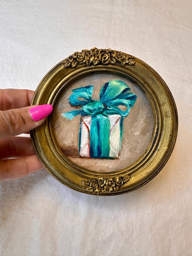 May include: A small circular painting of a white gift box wrapped with a vibrant turquoise ribbon and bow. The artwork is encased in an ornate, antique-style gold frame featuring sculpted floral details at the top and bottom edges.