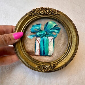 May include: A small circular painting of a white gift box wrapped with a vibrant turquoise ribbon and bow. The artwork is encased in an ornate, antique-style gold frame featuring sculpted floral details at the top and bottom edges.