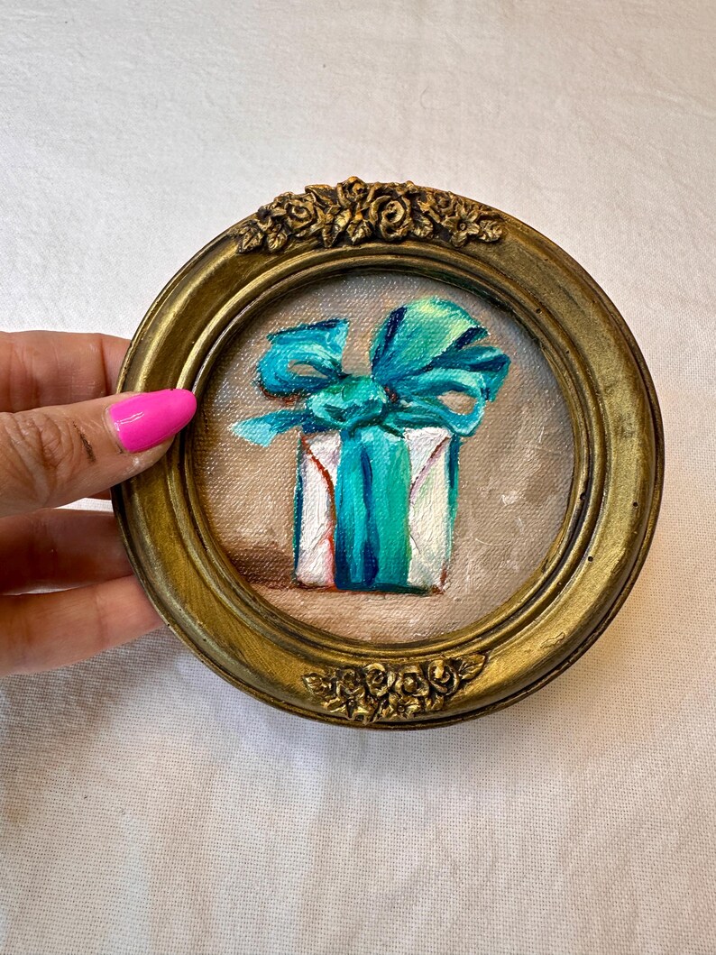 May include: A small circular painting of a white gift box wrapped with a vibrant turquoise ribbon and bow. The artwork is encased in an ornate, antique-style gold frame featuring sculpted floral details at the top and bottom edges.