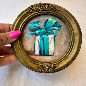 May include: A small circular painting in a gold-toned ornate frame featuring a white gift box tied with a vibrant turquoise ribbon. The artwork is rendered on canvas with visible brushstrokes, showcasing a central bow and a soft neutral background.