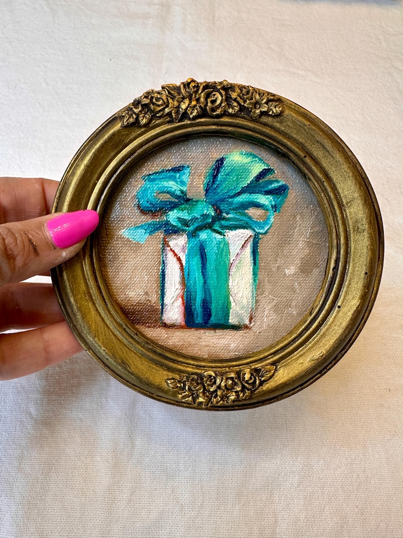May include: A small circular painting in a gold-toned ornate frame featuring a white gift box tied with a vibrant turquoise ribbon. The artwork is rendered on canvas with visible brushstrokes, showcasing a central bow and a soft neutral background.