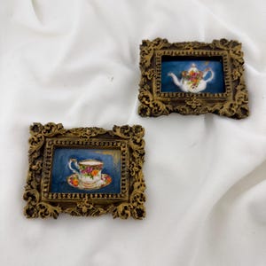 May include: Two small rectangular wall art pieces featuring ornate, antique-style gold frames. One frame displays a painting of a white teapot with floral patterns on a dark blue background, while the other shows a teacup and saucer with similar floral details.
