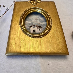 May include: A rectangular gold-toned picture frame with a central oval opening containing a small oil painting of a white house under a cloudy sky. The frame features a textured metallic finish and a decorative grape cluster motif at the top center.