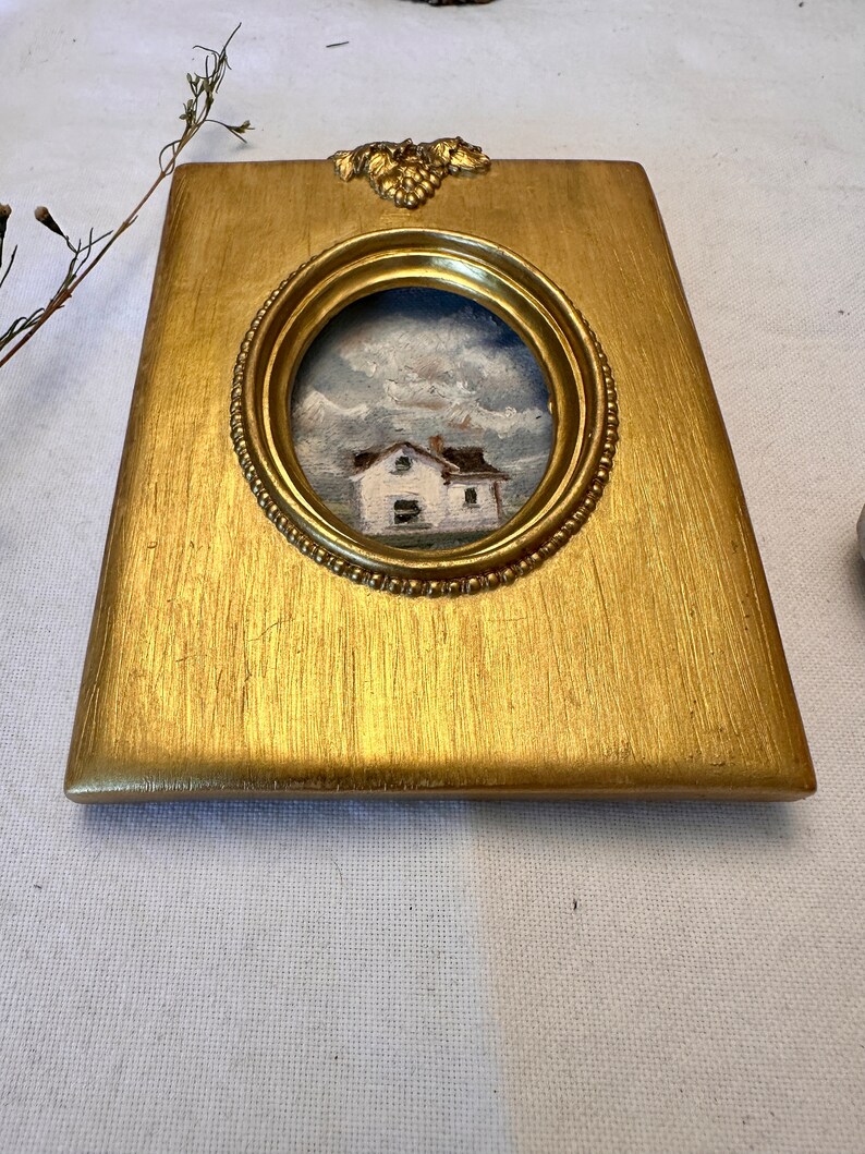 May include: A rectangular gold-toned picture frame with a central oval opening containing a small oil painting of a white house under a cloudy sky. The frame features a textured metallic finish and a decorative grape cluster motif at the top center.