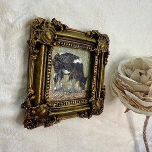 May include: A small square painting of a black and white cow grazing in a field, encased in an ornate, antique-style gold frame with intricate relief detailing. The artwork is positioned next to a large, pale, layered fabric flower.