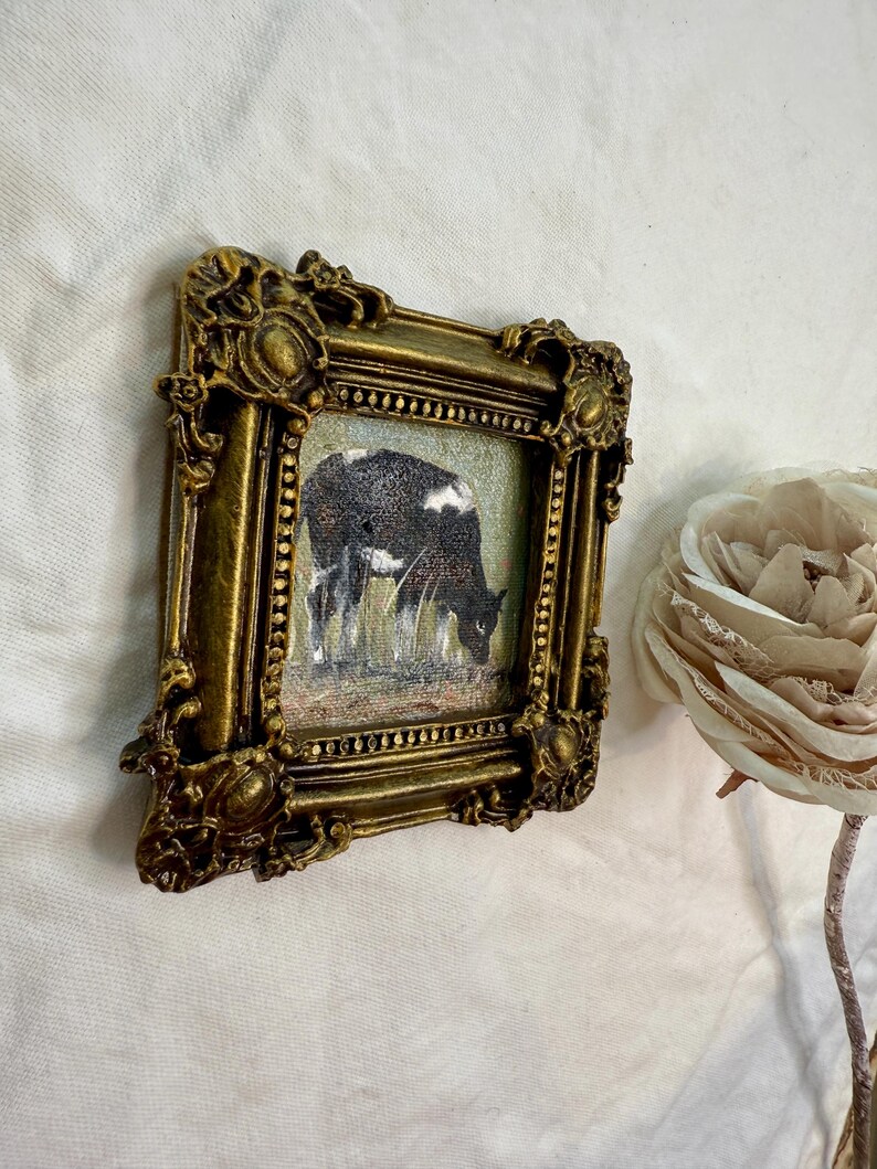 May include: A small square painting of a black and white cow grazing in a field, encased in an ornate, antique-style gold frame with intricate relief detailing. The artwork is positioned next to a large, pale, layered fabric flower.