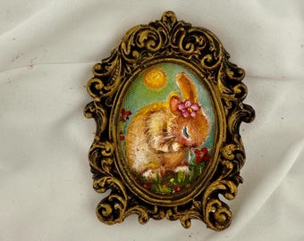 May include: An oval painting of a brown rabbit with a pink flower on its head, set against a green background with a yellow sun and red flowers. The artwork is encased in an ornate, antique-style gold frame with intricate scrollwork details.