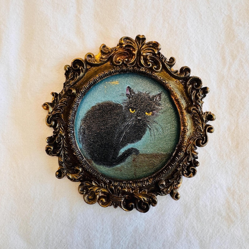May include: An antique-style framed picture of a black cat with yellow eyes on a light blue backdrop. The oval frame is gold-toned with elaborate detailing, giving a classic, vintage appearance. The frame is approximately 10 cm in diameter.